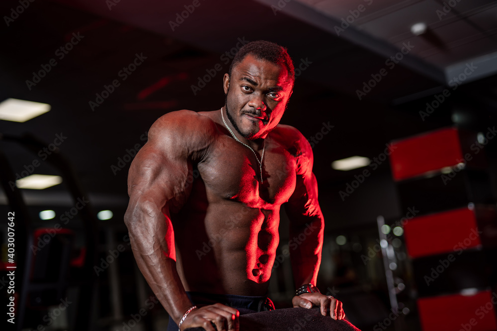 Portrait of an african american male fitness trainer. Bodybuilder shows biceps and looks at camera on gym background.