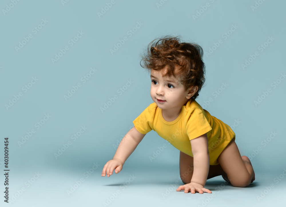 Cute smiling curly baby boy toddler in yellow comfortable jumpsuit crawling on floor and feeling cheerful over blue wall background. Happy childhood, stylish look for newborn and children Cute smiling curly baby boy toddler in yellow comfortable jumpsuit crawling on floor and feeling cheerful over blue wall background. Happy childhood, stylish look for newborn and children