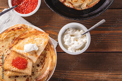 Traditional Russian Crepes Blini stacked in plate and cast-iron frying pan with red caviar, fresh sour cream on dark wooden table. Russian festival meal Maslenitsa or Shrovetide. Top view.