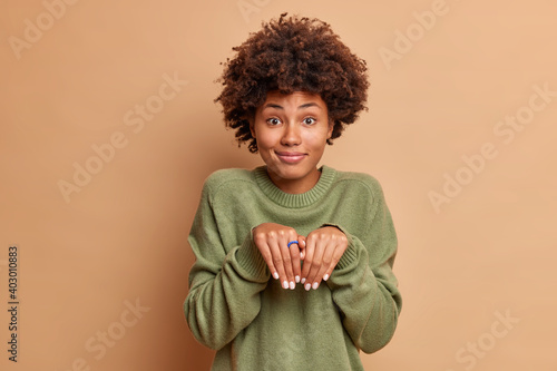 Playful pretty dark skinned African American woman makes bunny paws smiles pleasantly plays with child has fun wears long sleeved jumper poses against brown background. Female model turned into puppy