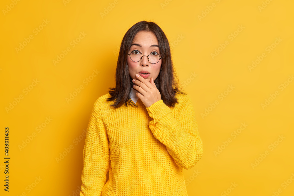 Impressed beautiful Asian woman holds chin looks stunned at camera hears something shocking wears round spectacles casual sweater isolated over yellow background. People and reaction concept Impressed beautiful Asian woman holds chin looks stunned at camera hears something shocking wears round spectacles casual sweater isolated over yellow background. People and reaction concept