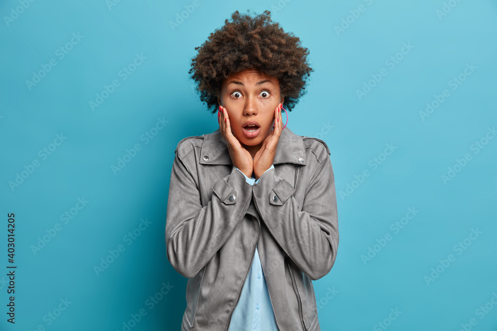 Photo of emotional dark skinned woman grabs face stares with eyes full of fear cannot believe in shocking news has curly bushy hair wears grey jacket isolated over blue background. Omg concept Photo of emotional dark skinned woman grabs face stares with eyes full of fear cannot believe in shocking news has curly bushy hair wears grey jacket isolated over blue background. Omg concept