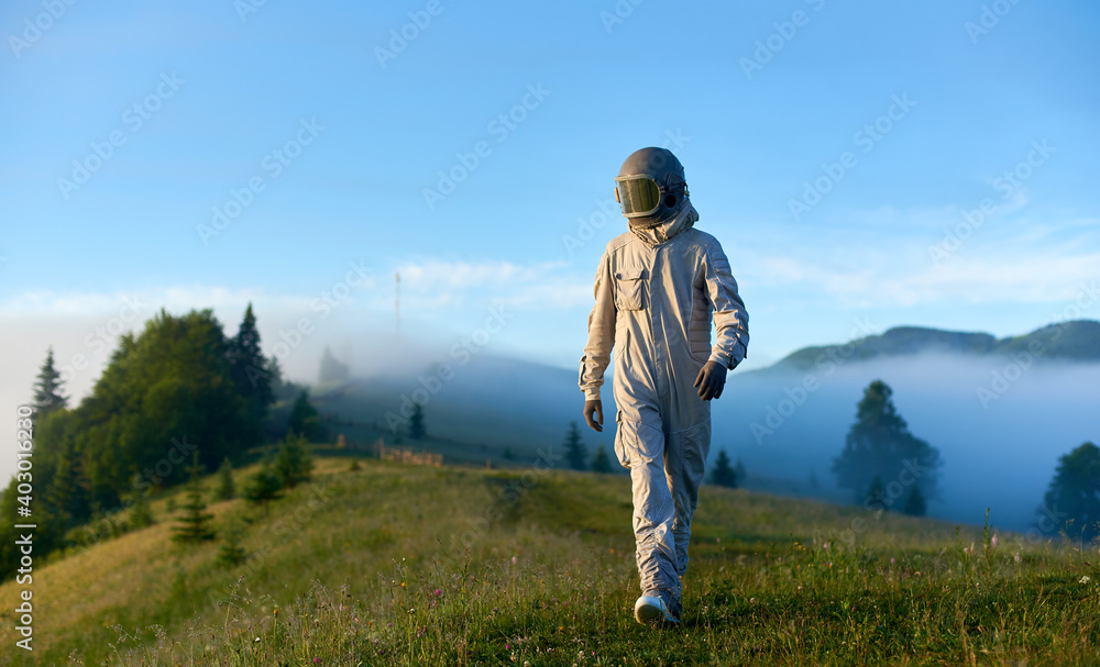Spaceman wearing white space suit and helmet walking alone sunny green mountain glade in the morning, foggy hills and blue sky on background. Concept of astronautics, Earth exploration and nature