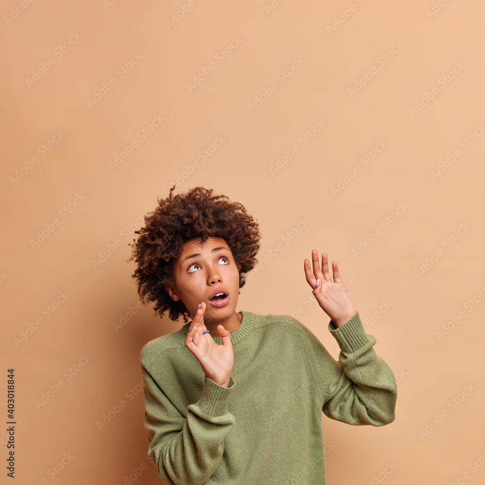 Frightened African American woman keeps arms raised tries to stop something falling above concentrated upwards being afraid wears casual jumper poses against beige background copy space area Frightened African American woman keeps arms raised tries to stop something falling above concentrated upwards being afraid wears casual jumper poses against beige background copy space area