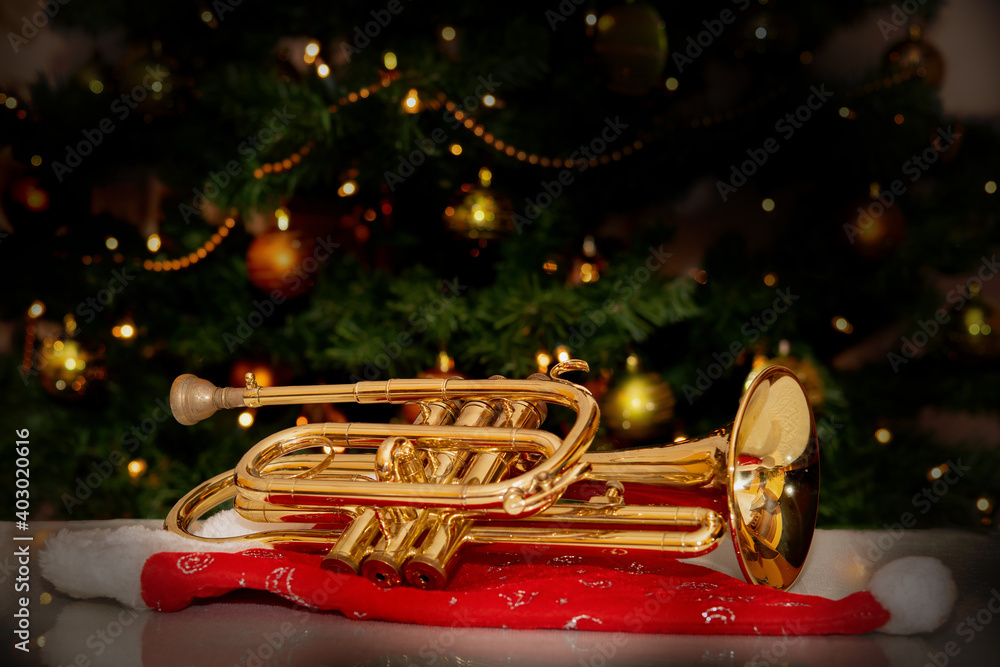 Cornet – brass musical instrument – placed on Santa’s hat with a Christmas tree in the background. Festive holiday atmosphere with warm lights and musical charm.