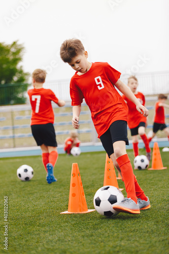 Boys kicking football balls on outdoor training. Group of young soccer players practicing dribbling skills running balls between training cones. Youth soccer team in red jersey shirts