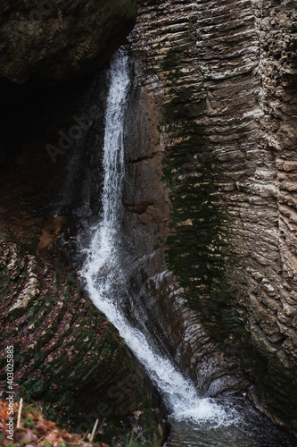 Walk by river in gorge and view beautiful stormy waterfall. Small narrow waterfall in national park. Water falls from cliff and breaks down on surface of river and rocks.