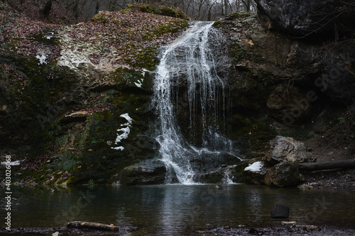 Walk by river in gorge and view beautiful stormy waterfall. Small narrow waterfall in national park. Water falls from cliff and breaks down on surface of river and rocks.