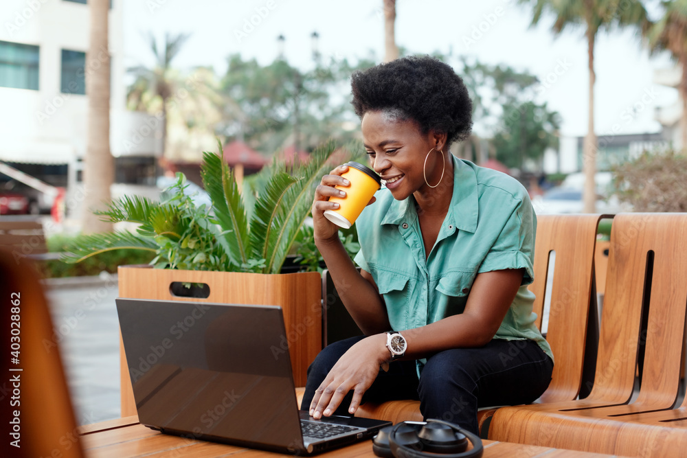 Fototapeta premium A beautiful African woman drinks coffee in a cafe and works at a laptop, headphones and a phone are lying next to her. The girl works at the computer during a coffee break