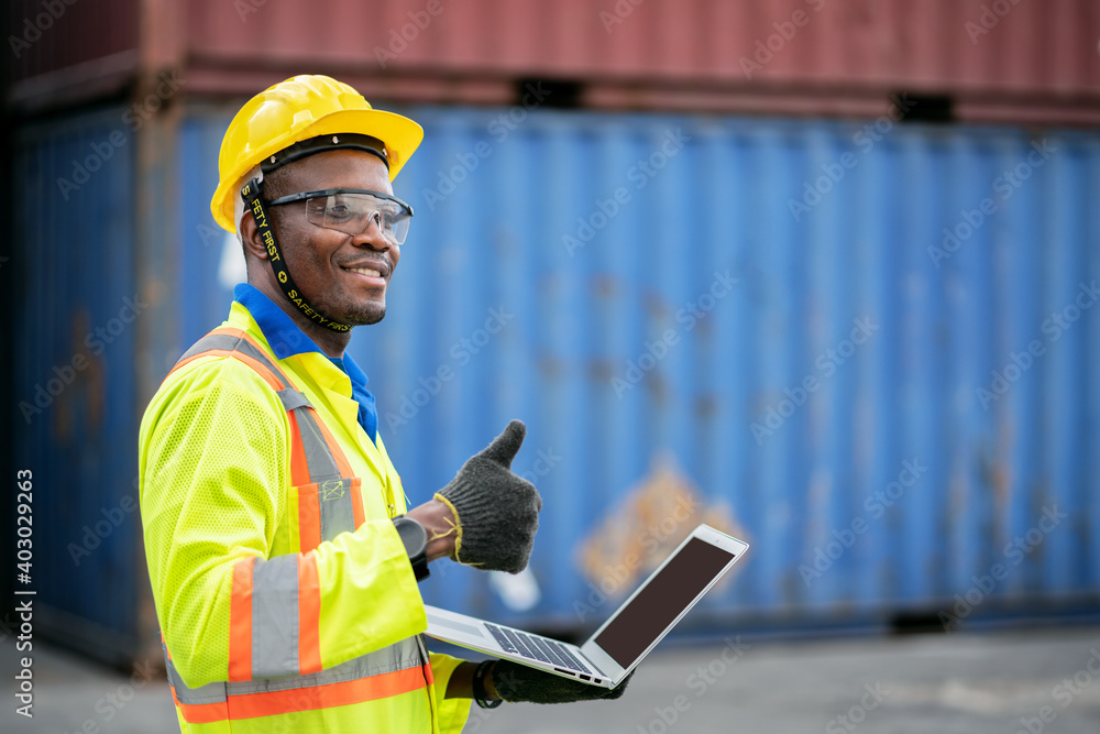 Portrait of African happy worker in protective safety jumpsuit uniform with hardhat and use laptop computer at cargo container shipping warehouse. transportation import,export logistic industrial Portrait of African happy worker in protective safety jumpsuit uniform with hardhat and use laptop computer at cargo container shipping warehouse. transportation import,export logistic industrial