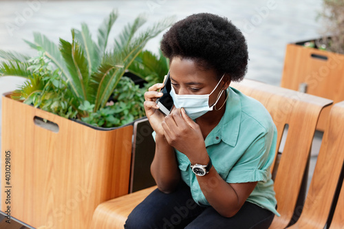 A young African woman wearing a medical face mask talks on the phone while sitting on a bench. Social distancing and work, work online, business online