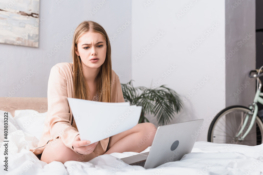 young freelancer in silk robe looking at documents near laptop on bed young freelancer in silk robe looking at documents near laptop on bed