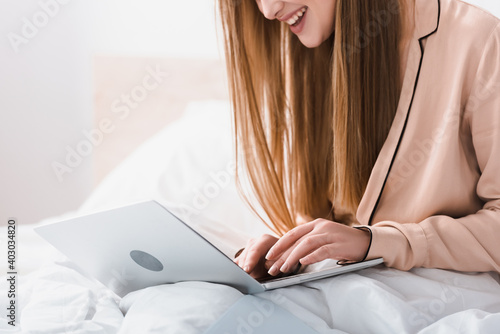 cropped view of smiling young freelancer in silk robe using laptop in bedroom