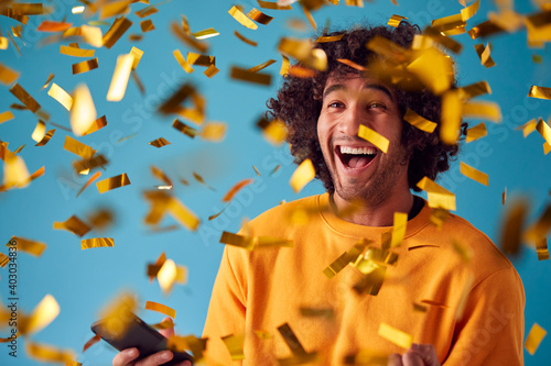 Celebrating Young Man With Mobile Phone Winning Prize And Showered With Gold Confetti In Studio photo