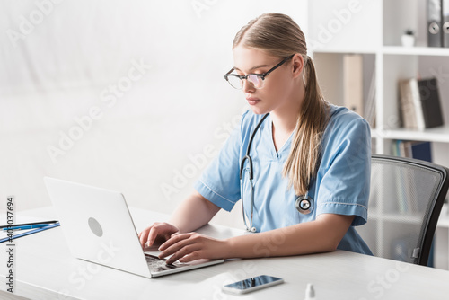 young veterinarian in glasses typing on laptop near smartphone on desk