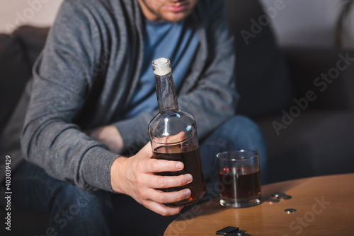 partial view of lonely man holding bottle of whiskey near glass, blurred background
