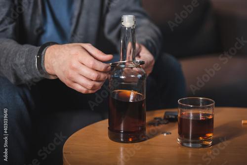 cropped view of man, handcuffed to bottle of whiskey, sitting at home alone
