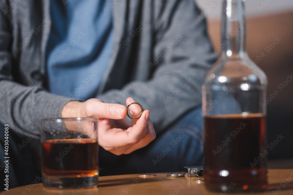 selective focus of wedding ring in hand of alcoholic man sitting near glass and bottle of whiskey
