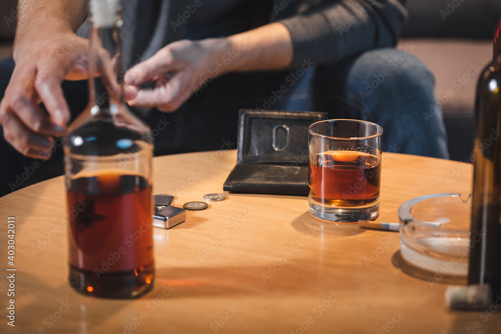cropped view of alcohol-addicted man taking bottle of whiskey from table near empty wallet, blurred foreground cropped view of alcohol-addicted man taking bottle of whiskey from table near empty wallet, blurred foreground