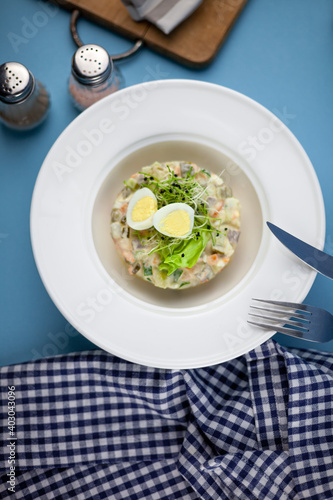 Traditional salad Olivier. Christmas and New year food. White plat with fork and knife on blue background with copy space. Top view, flat lay. Microgreen, quail eggs. Serving at a restaurant.
