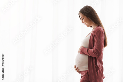 Portrait Of Smiling Pregnant Woman Tenderly Embracing Belly Against Window At Home