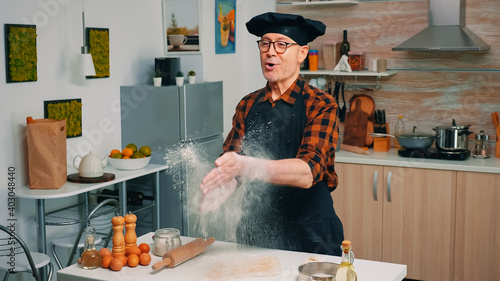 Baker wearing a uniform throws up flour standing near the table. Professional cook with bonete claps and powder fly, retired chef sprinkling rew ingredients over dough preparing traditional bread. photo