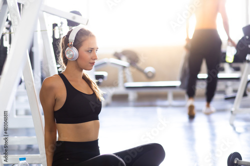 Picture of attractive fitness woman sitting on the floor at gym while using phone and listening music with headphones