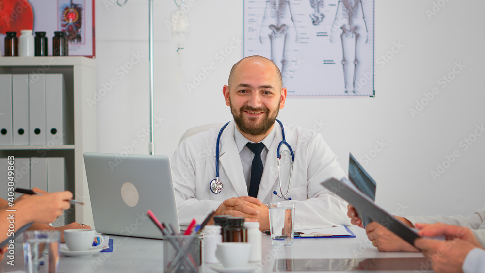 Portrait of doctor typing on laptop during brainstorming and looking at camera smiling sitting on desk in hospital office, conference room. Specialist in lab coat writing on notepad in medical clinic. Portrait of doctor typing on laptop during brainstorming and looking at camera smiling sitting on desk in hospital office, conference room. Specialist in lab coat writing on notepad in medical clinic.