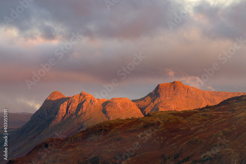 Beautiful red sunlight from the rising sun illuminating the Langdale Pikes mountain range in the English Lake District with dark clouds in sky.