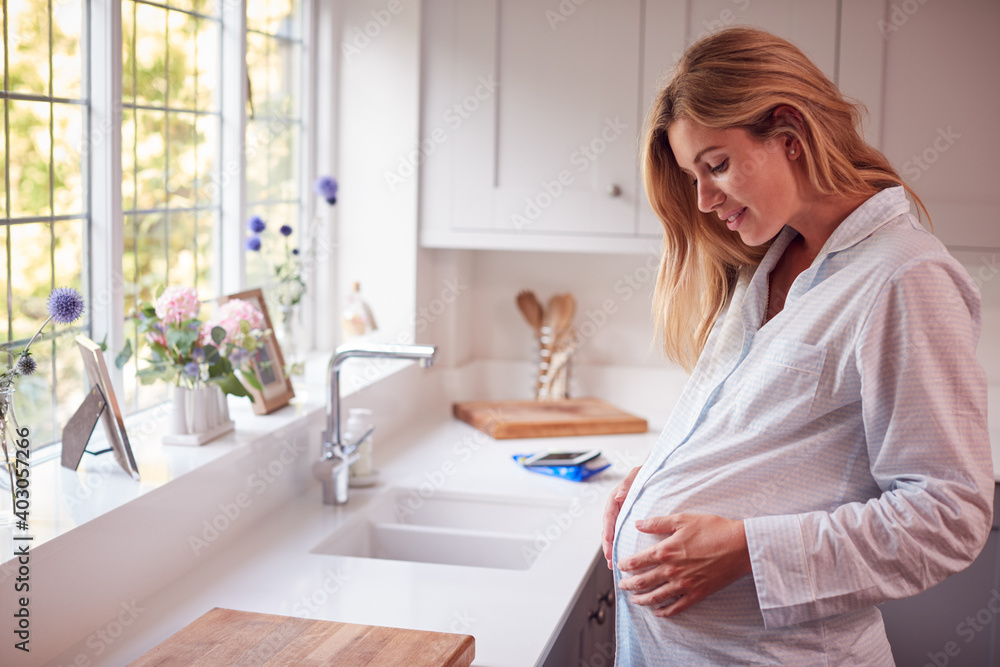 Pregnant Woman Wearing Pyjamas Standing In Kitchen Holding Bump - obrazy, fototapety, plakaty 
