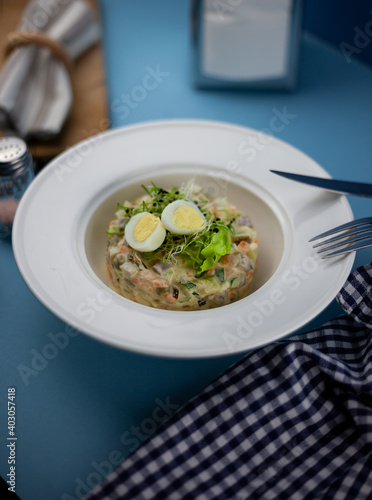 Traditional salad Olivier. Christmas and New year food. White plat with fork and knife on blue background with copy space. Top view, flat lay. Microgreen, quail eggs. Serving at a restaurant.