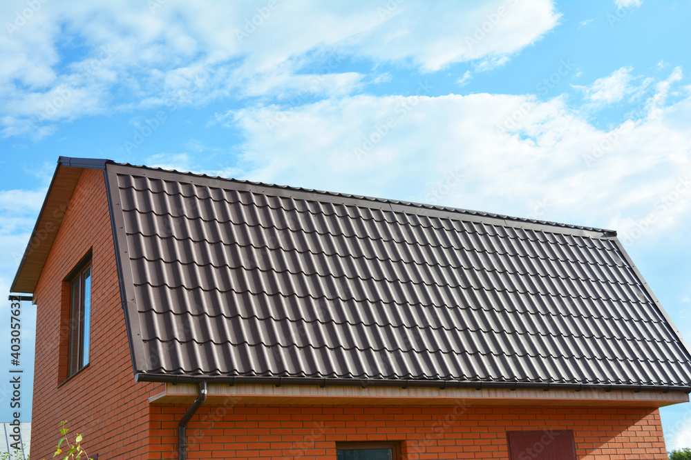 Fototapeta premium A close-up of a mansard metal roof with an attic window and roof gutters of a brick house construction against the blue sky.