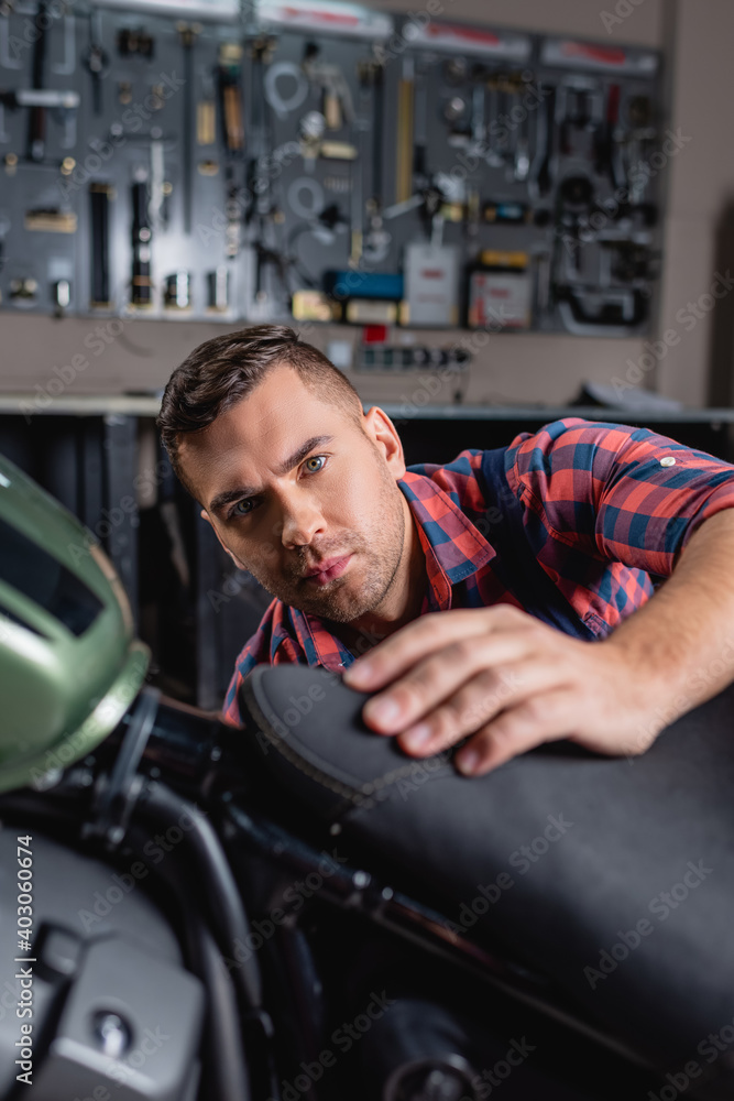 young mechanic looking at camera while making diagnostics of motorbike on blurred foreground