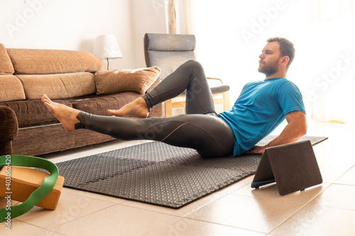 Fotografia young man doing crunches at home on sports mat with raised legs, bright room with tablet and yoga blocks around