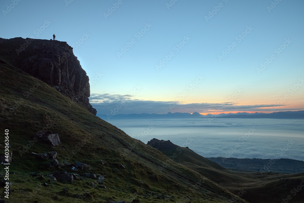 Silhouette of a traveler standing on a large rock and looking at the sea before the rising sun. Isle of Skye, Scotland