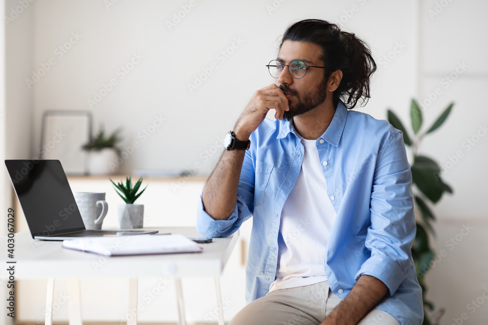 Thoughtful young arab male freelancer sitting at desk at home, looking aside Thoughtful young arab male freelancer sitting at desk at home, looking aside
