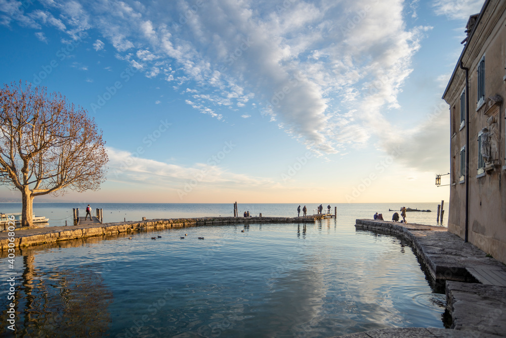 Scenic sunset at Punta San Vigilio, popular Italian touristic destination on Garda Lake, Lombardy. Clouds in the sky reflect in the water, people silhouette enjoying the sunset Scenic sunset at Punta San Vigilio, popular Italian touristic destination on Garda Lake, Lombardy. Clouds in the sky reflect in the water, people silhouette enjoying the sunset