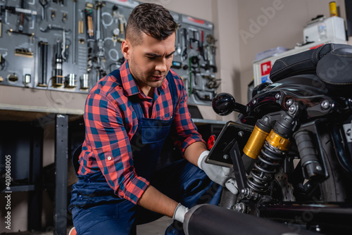 young mechanic in overalls and plaid shirt checking shock absorber of motorcycle in workshop
