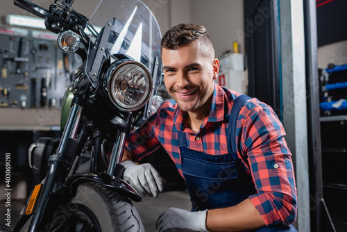 cheerful mechanic in overalls smiling at camera near motorbike in workshop