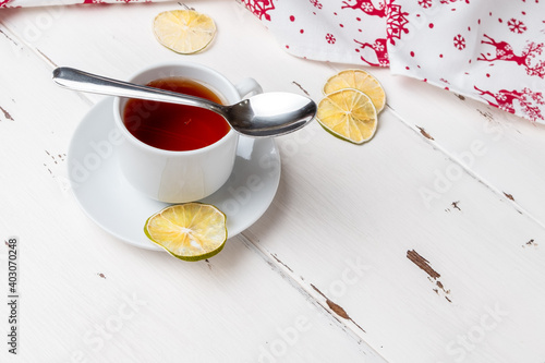 White ceramic cup with tea on a saucer. Dried lime slices. Napkin with Christmas ornament. New Year and Christmas decor on a white wooden background. Copy space.