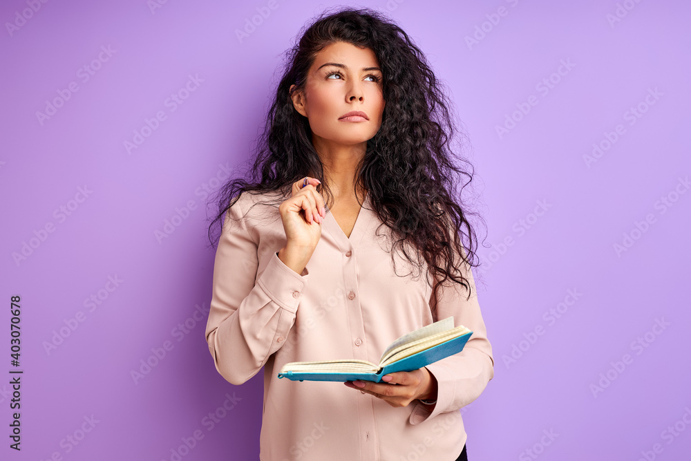 curly woman stand thinking before making notes in notebook, isolated over purple background curly woman stand thinking before making notes in notebook, isolated over purple background