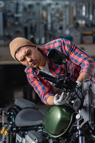 young mechanic in plaid shirt and beanie checking motorcycle handlebar in workshop