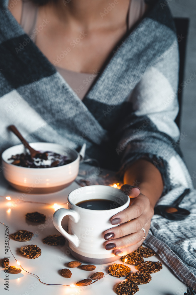 The woman is wrapped in a blanket and holds a cup of coffee in her hands. Winter comfort, garlands The woman is wrapped in a blanket and holds a cup of coffee in her hands. Winter comfort, garlands