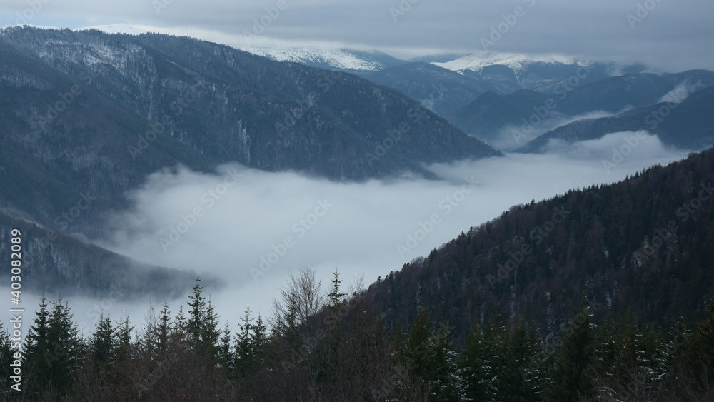 Clouded winter scenery. A gray fog covers the low lands forests and valleys. The snow covers the high altitude crests of the mountain peaks. Parang, Carpathia, Romania.