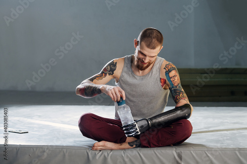 Cyborg man with bionic hand sitting on a mat, opening plastic bottle with water, taking a break. He is looking down, tired. Beautiful tattoos on shoulders. Frontal view.