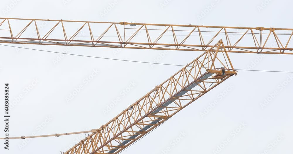 Two turning yellow tower crane jibs on a construction site against white cloudy sky Two turning yellow tower crane jibs on a construction site against white cloudy sky