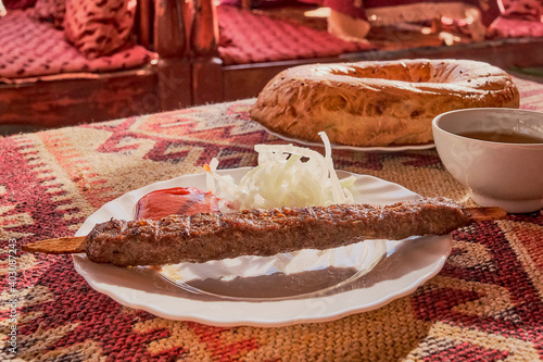 Lamb lula kebab with tomato sauce and pickled onions on a white plate. On the table is a tablecloth with a red asian pattern. photo