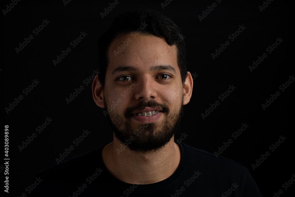 Young man frontal head shot with black background. He is smiling. He has a casual look with beard and t-shirt.
