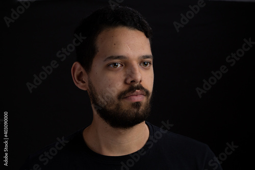 Young man head shot with black background. He is serious. He has a casual look with beard and t-shirt.