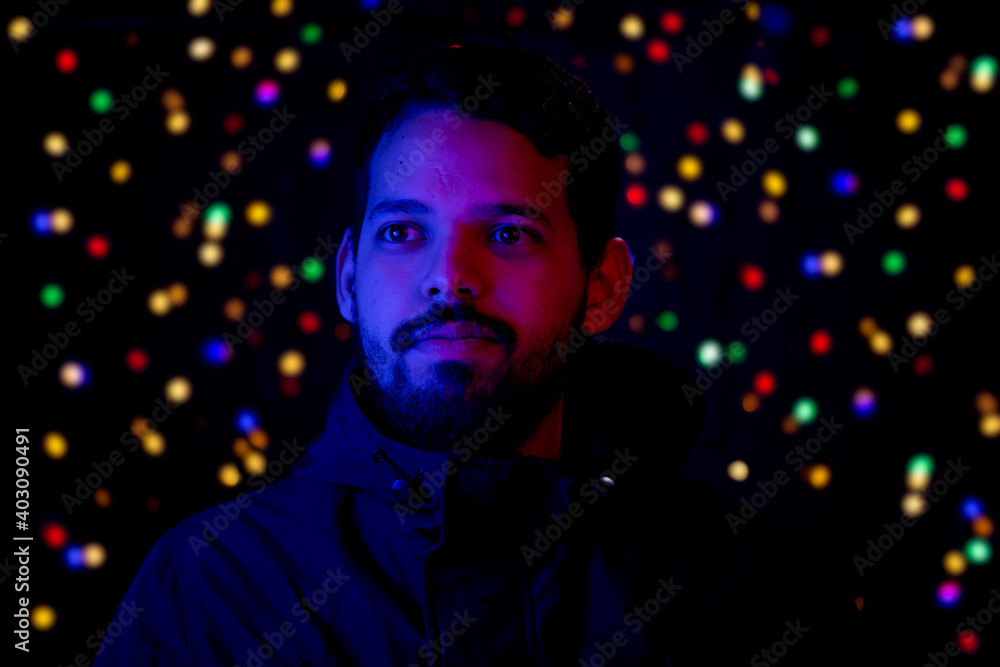 Young man head shot illuminated with red and blue lights on face and holiday lights as background. He is smiling. He has a casual look with beard and winter jacket.